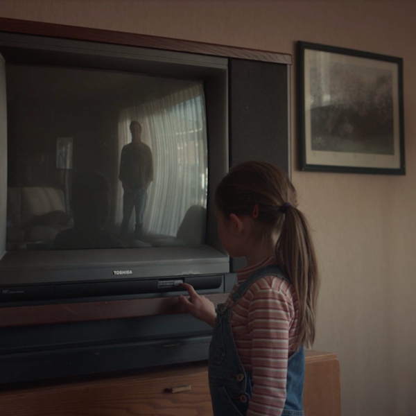 A little girl with her hand on the power button of a tv and the reflection of her father in the tv.